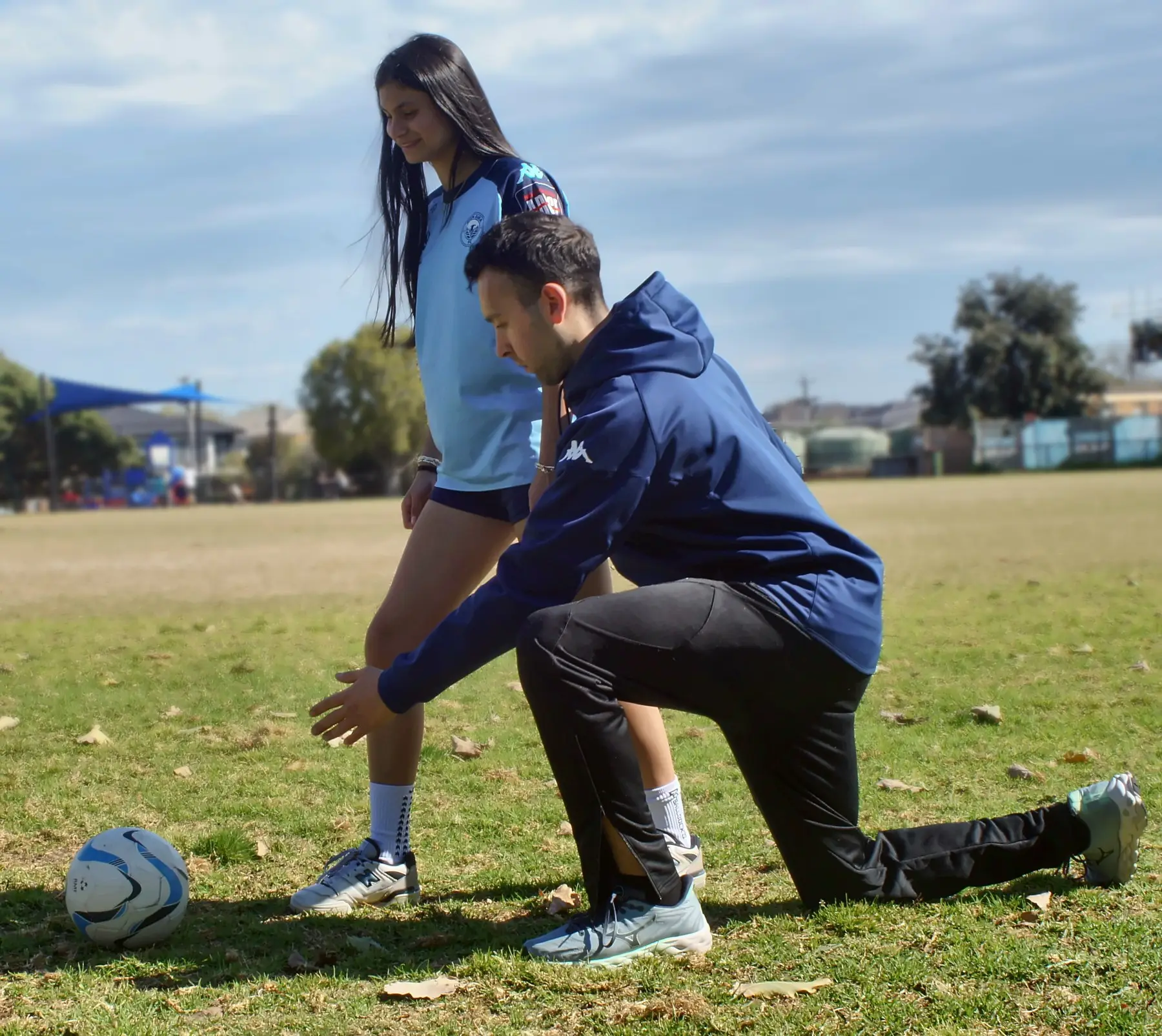 Jake kneeling beside a young female footballer, coaching her knee and foot position on the field